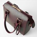 Maroon structured top handle handbag - model close-up holding bag showing gold chain charm and flower tab